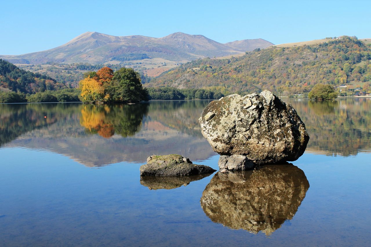 Explore the serene autumn scenery of Chambon Lake with colorful reflections and distant mountains.